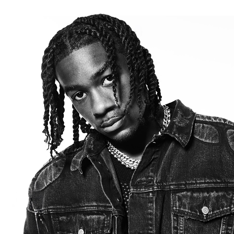 Black and white portrait of a young man with twisted hair, wearing a denim jacket and layered necklaces, looking directly at the camera.