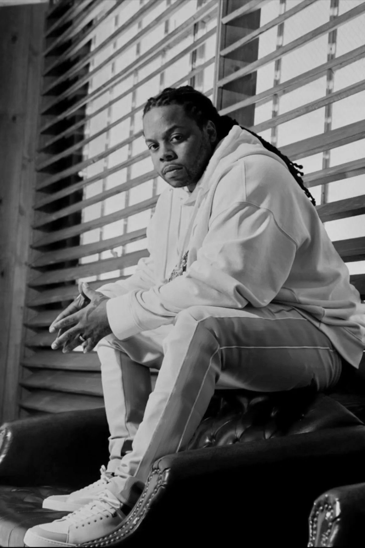 A man with dreadlocks wearing a white hoodie and light-colored pants, sitting on a leather armchair beside a wooden slat wall, looking at the camera in black and white.