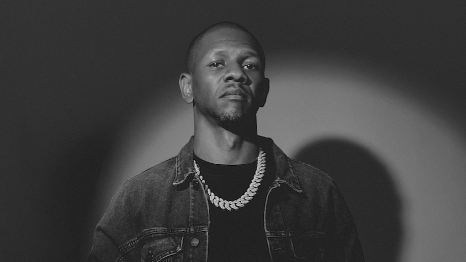 Black and white portrait of a young man wearing a denim jacket and a chain necklace, looking serious.
