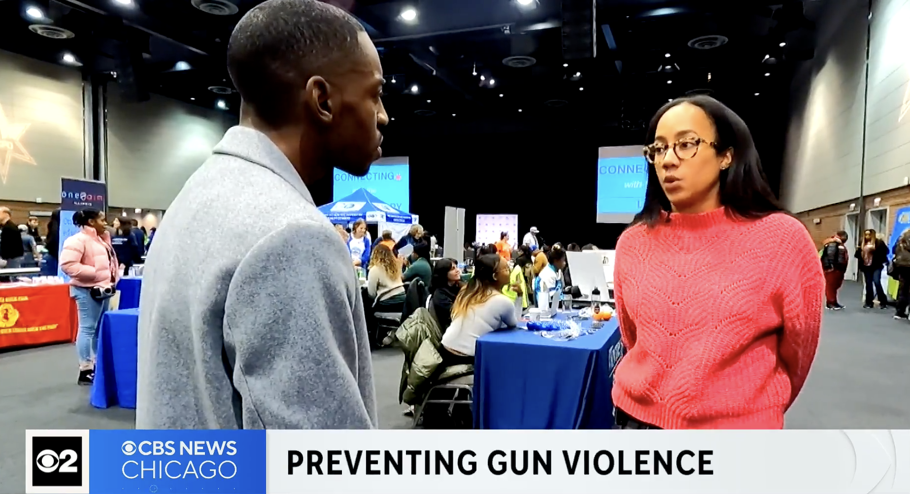 A woman in a pink sweater and glasses talks to a man in a gray jacket at a conference, with a large screen and people at tables in the background.