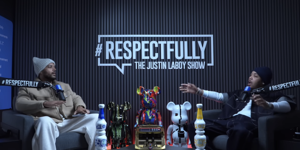 Two men sitting in a podcast studio, with a black wall and a sign that reads '#Respectfully The Justin Laboy Show'. They are surrounded by colorful bear-shaped sculptures and bottles, engaged in conversation with microphones.