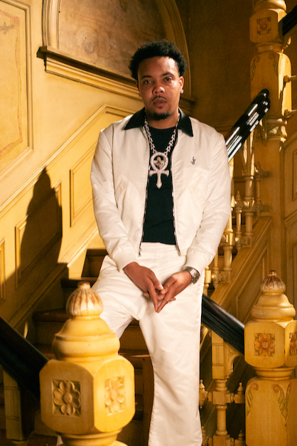 Man dressed in white jacket and pants, standing on a staircase in a vintage-style interior with wooden railings and detailed woodwork.