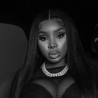 A young woman with long straight hair, wearing makeup, a mesh top, and a pearl necklace, posing for a photo in black and white.