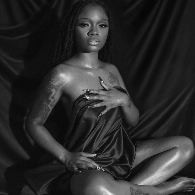 Black and white photo of a woman sitting on a dark fabric backdrop, topless and holding a silky dark cloth across her chest, with tattoos on her arms and chest, looking confidently at the camera.