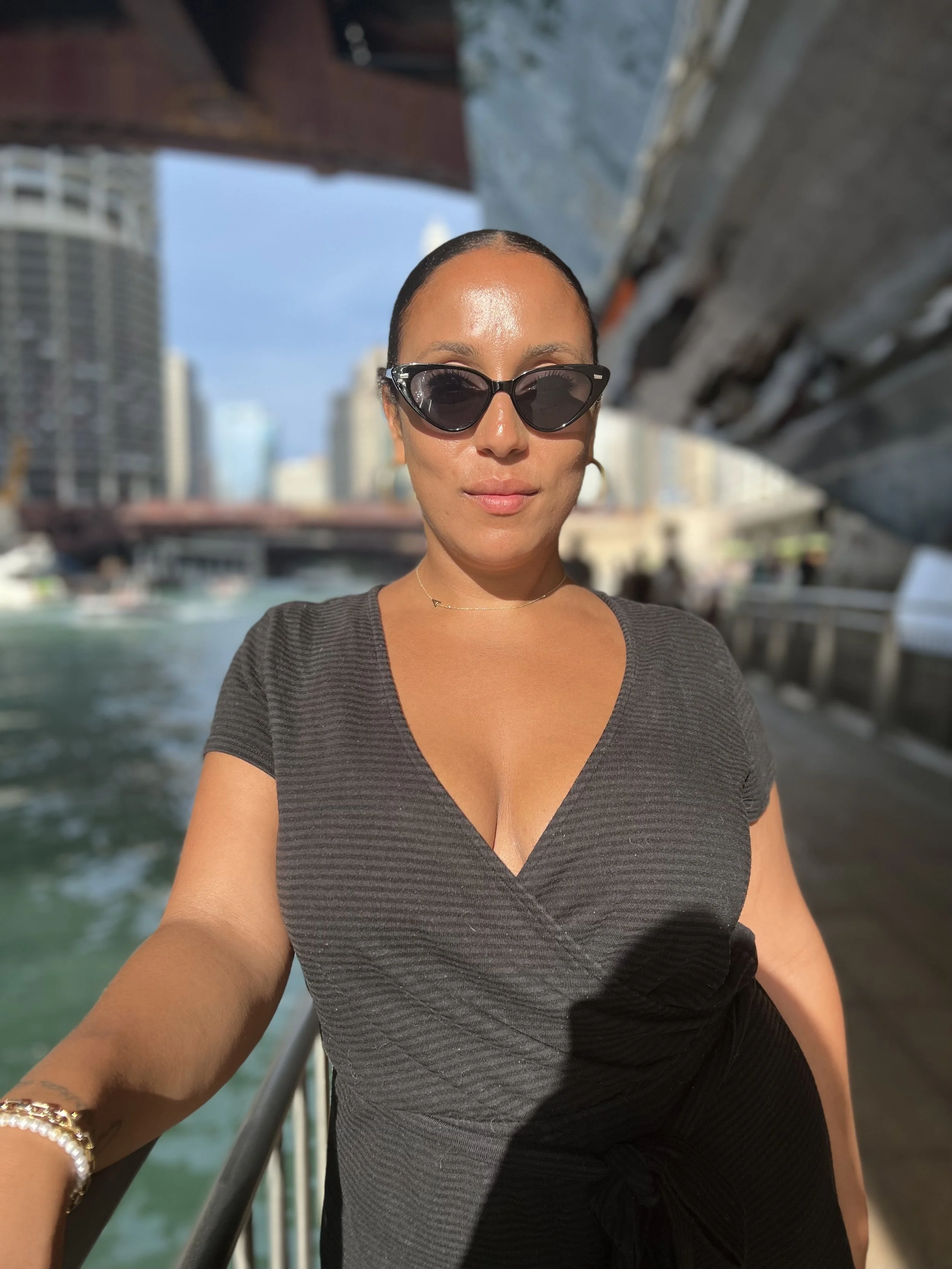 A woman taking a selfie outdoors with a city skyline and water in the background. She is wearing black sunglasses, a black dress with a V-neck, and gold jewelry.