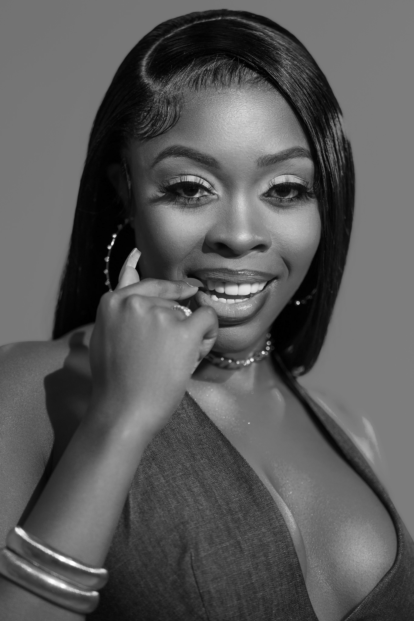 Portrait of a smiling woman with straight hair, wearing jewelry, touching her face, in black and white.