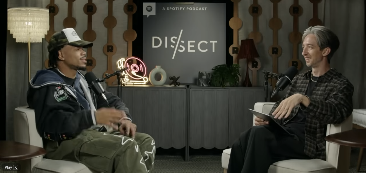 Two men sitting in a podcast studio having a conversation. The man on the left wears a camouflage baseball cap, jacket, and cargo pants. The man on the right has gray hair, wears a checkered jacket, and holds a tablet. Behind them are decorated walls with a Spotify Neon sign, a lamp, and a sign that says 'DISSECT'.