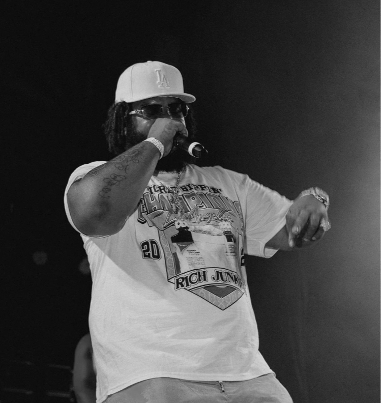 Man performing on stage, wearing a Los Angeles baseball cap, sunglasses, and a graphic T-shirt, holding a microphone.