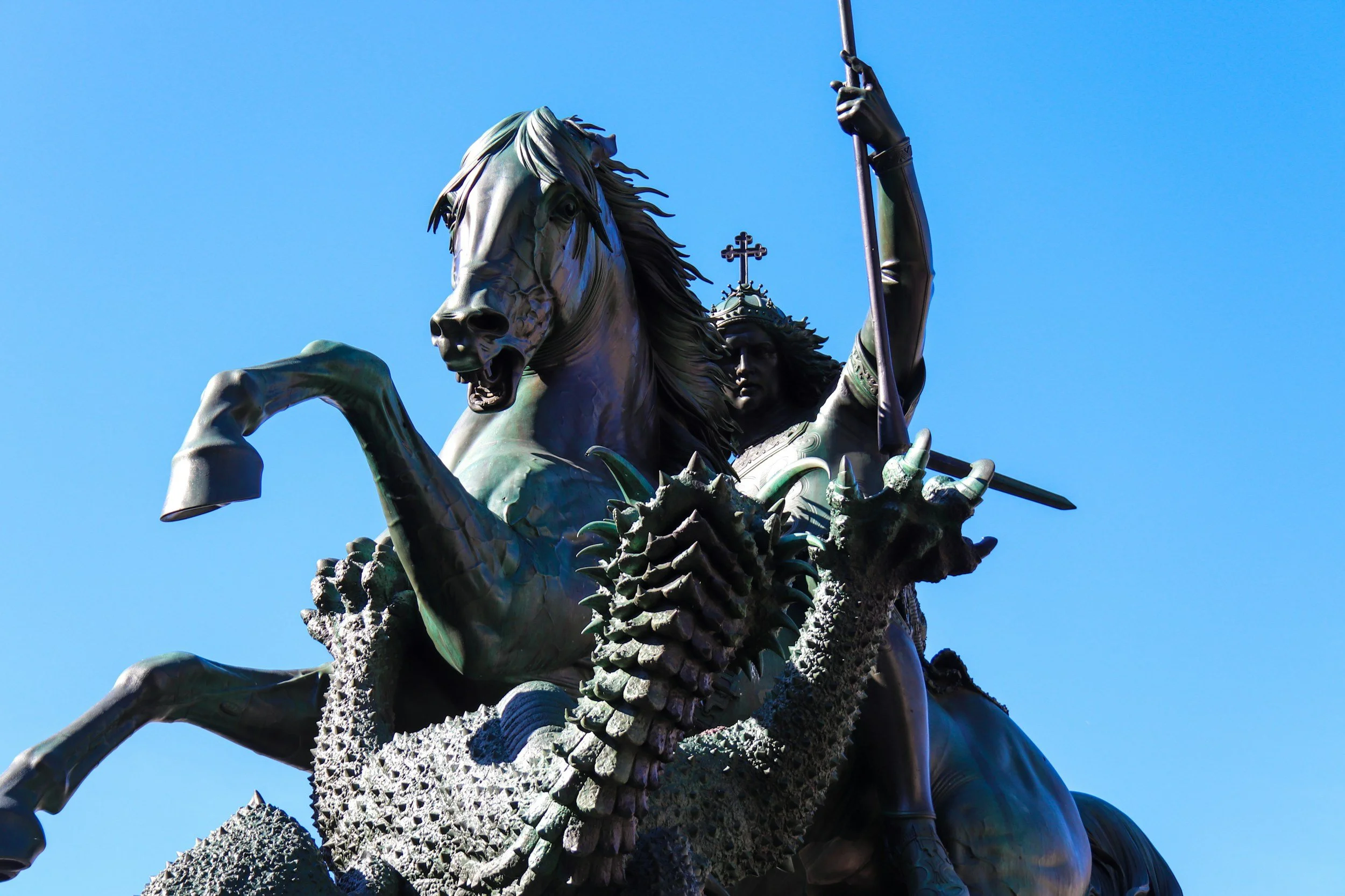 Bronze statue of a native American warrior riding a horse, holding a spear, with a dragon-like creature at the horse's front, against a clear blue sky.