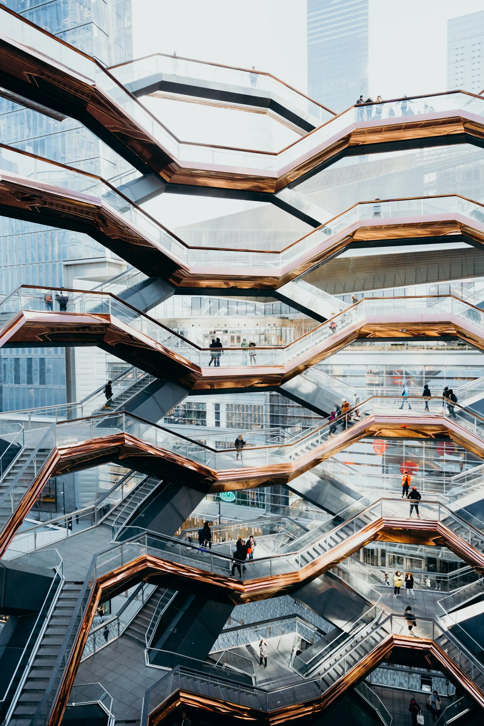 Multiple levels of outdoor walkways with people walking, set against tall modern glass buildings in an urban cityscape.