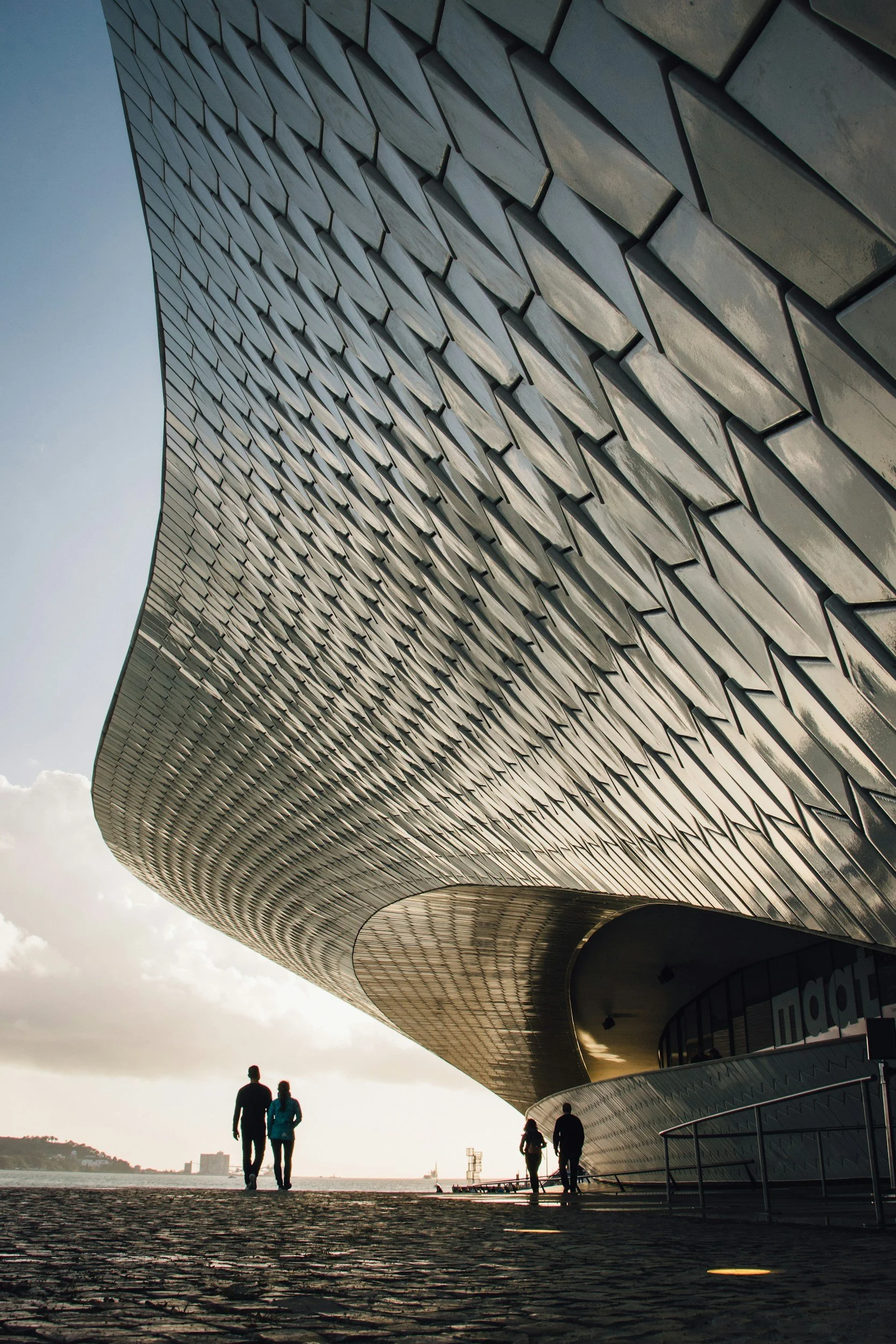 A modern building with a curved metallic facade and a textured pattern, with people walking nearby against a cloudy sky.