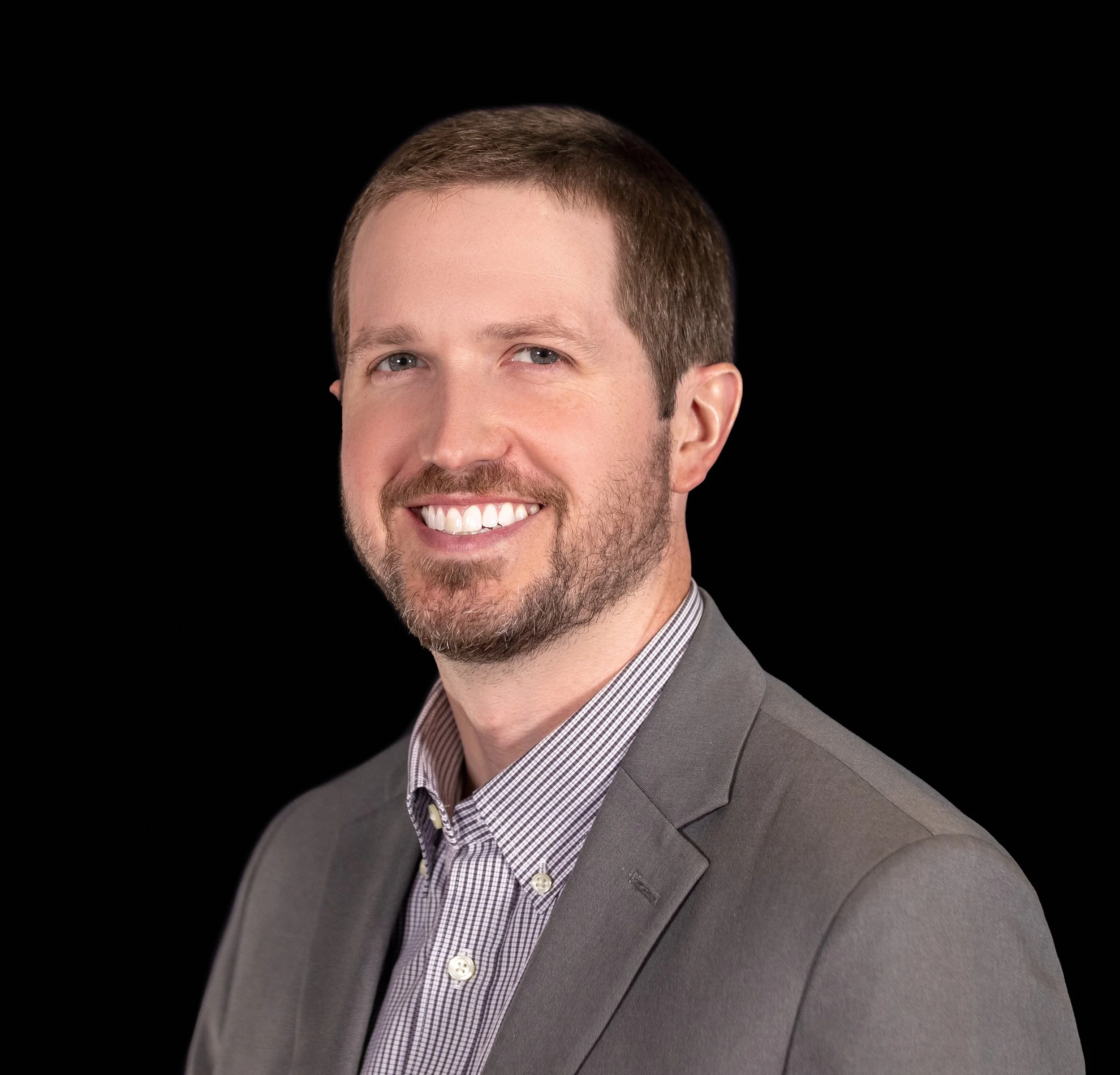 Portrait of a smiling man with short brown hair, beard, wearing a gray suit jacket and checkered dress shirt against a black background.