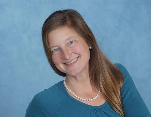Headshot of a woman with long brown hair, wearing a blue top and a pearl necklace, smiling against a blue background.