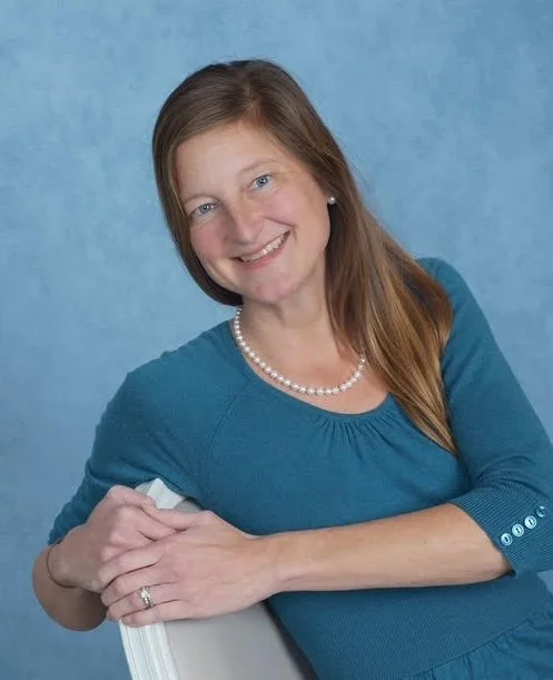 Karrisa Grellner: A woman with long brown hair, smiling, wearing a blue shirt, a necklace, and earrings, sitting and leaning on a chair with a blue background.