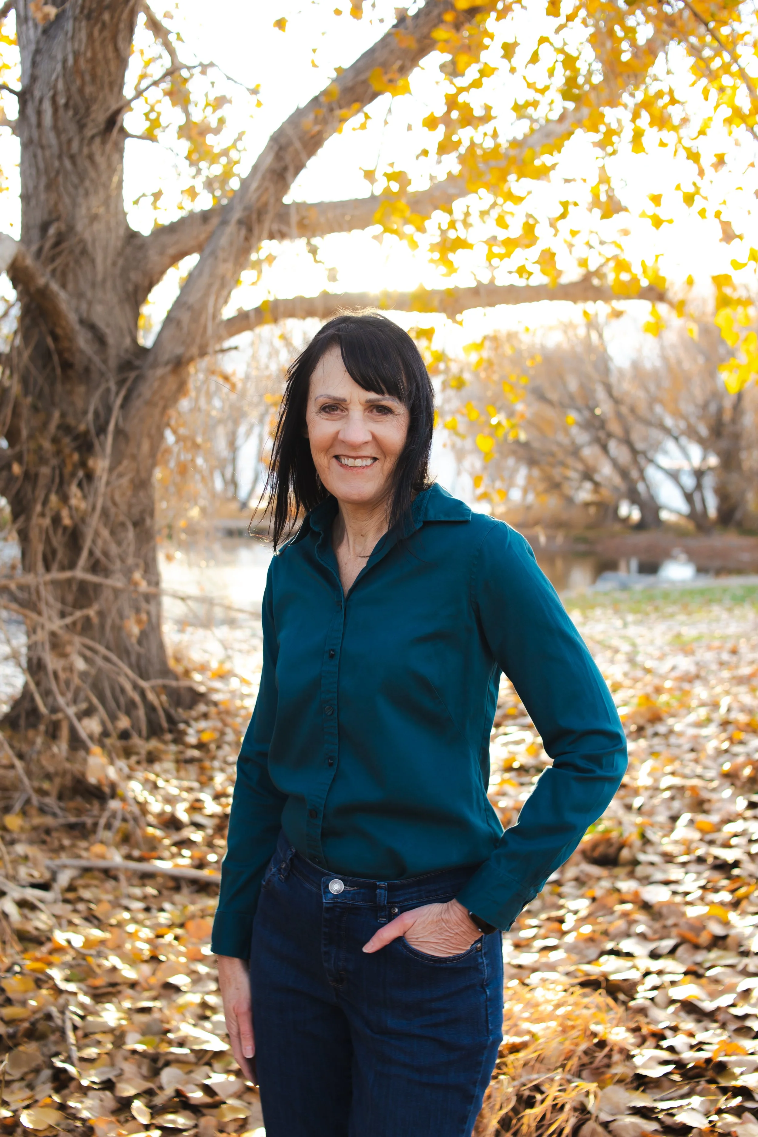 A picture of ClearSite Designer Alison in front of a pond surrounded by fall leaves. Alison wears a teal shirt and jeans