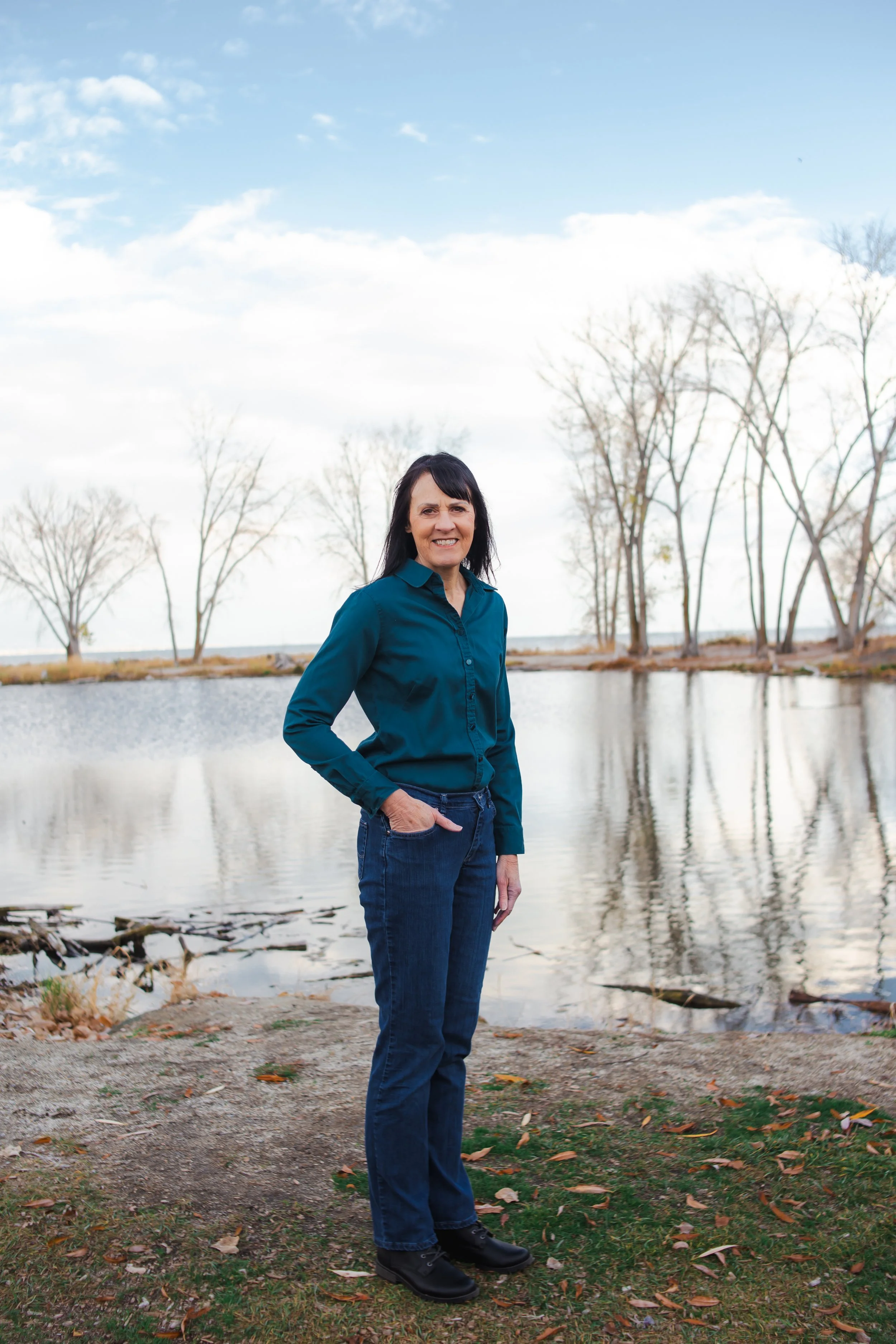 Alison, founder of ClearSite Design, stands in front of a pond which is backed by a lake. She wears a turquoise button-down and jeans.
