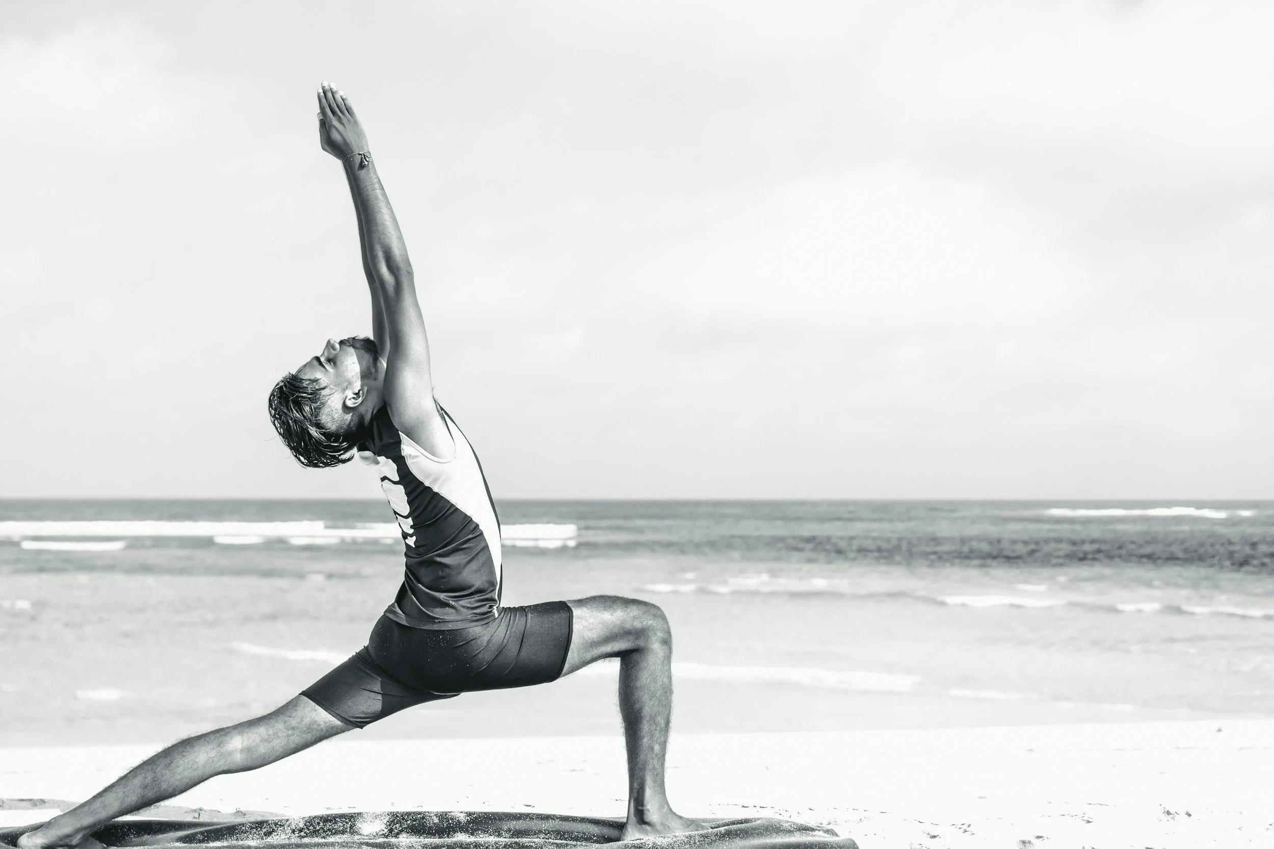 A man practicing yoga on the beach, performing a low lunge pose with arms raised overhead, near the ocean.