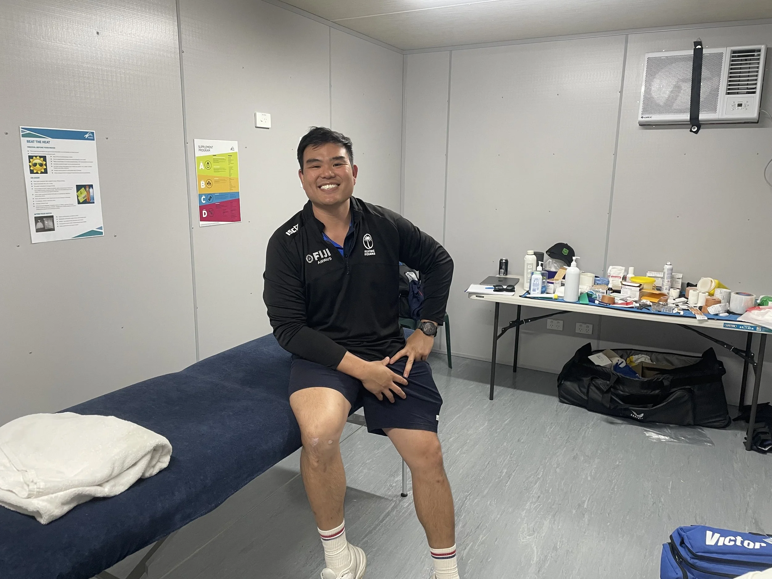Smiling man sitting on a blue examination table in a medical room with a towel and a large table filled with medical supplies in the background.