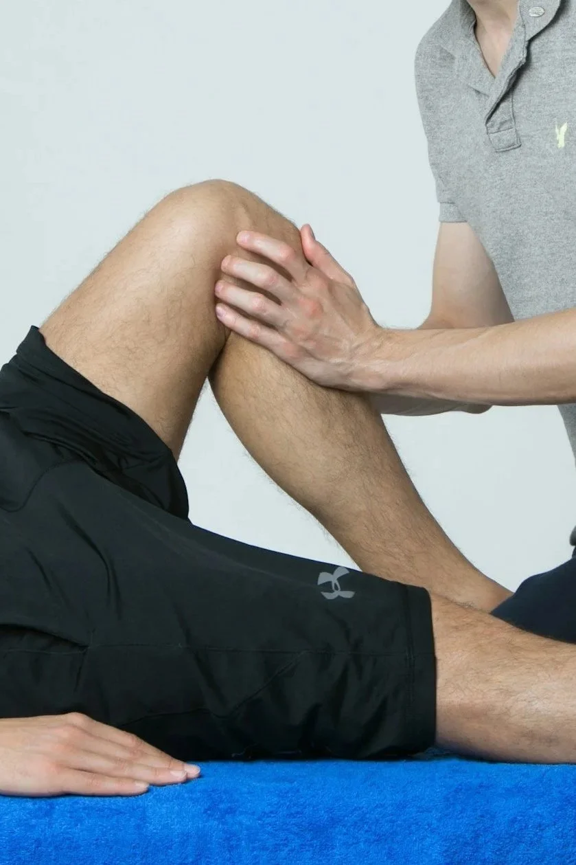 A person receiving knee therapy or physical therapy from a healthcare professional. The person is seated on a blue mat, wearing black shorts, and the healthcare provider is holding their knee, which is slightly bent.