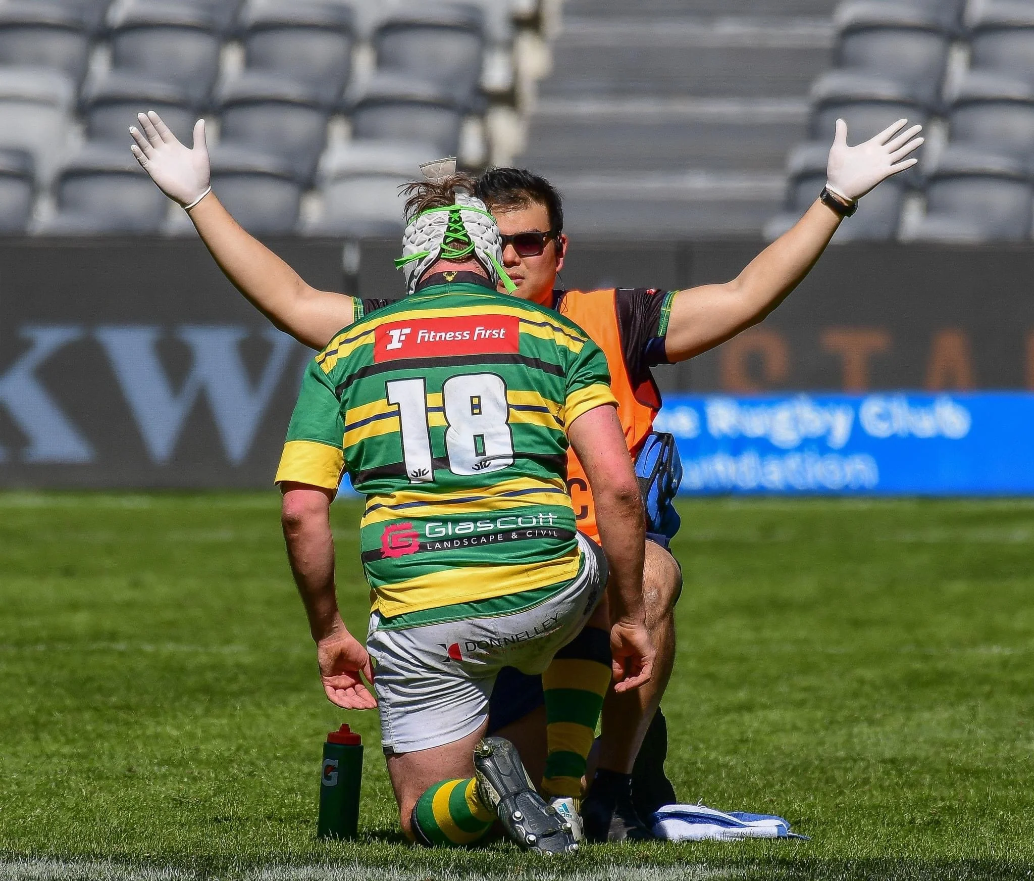 A rugby player wearing a green and yellow striped jersey with the number 18 is kneeling on the field, while a medic or trainer with outstretched arms stands behind him in a stadium. The rugby player appears to be receiving medical attention.