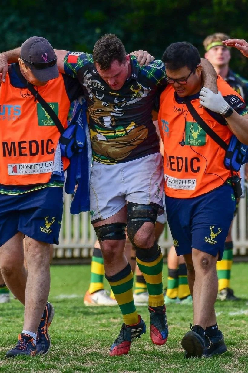 A rugby player with a knee injury being supported by two medics during a game.