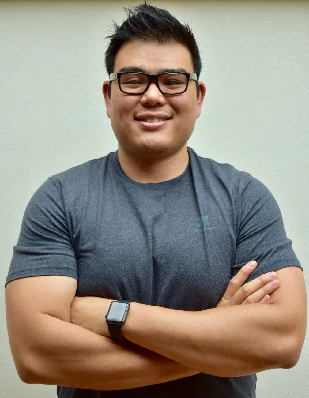 A young Asian man with short spiky black hair, glasses, and a smile, standing against a plain cream-colored wall with arms crossed. He is wearing a dark gray T-shirt and a black smartwatch on his left wrist.