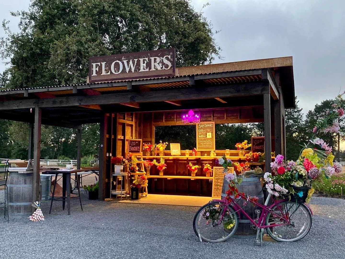 A flower stand at dusk with a pink bicycle decorated with flowers in a basket in front. The stand has a sign that reads 'FLOWERS' and is filled with colorful flower arrangements, some on shelves and others in buckets. There are chalkboard signs with prices and a purple light above the stand, set against trees and a cloudy sky.