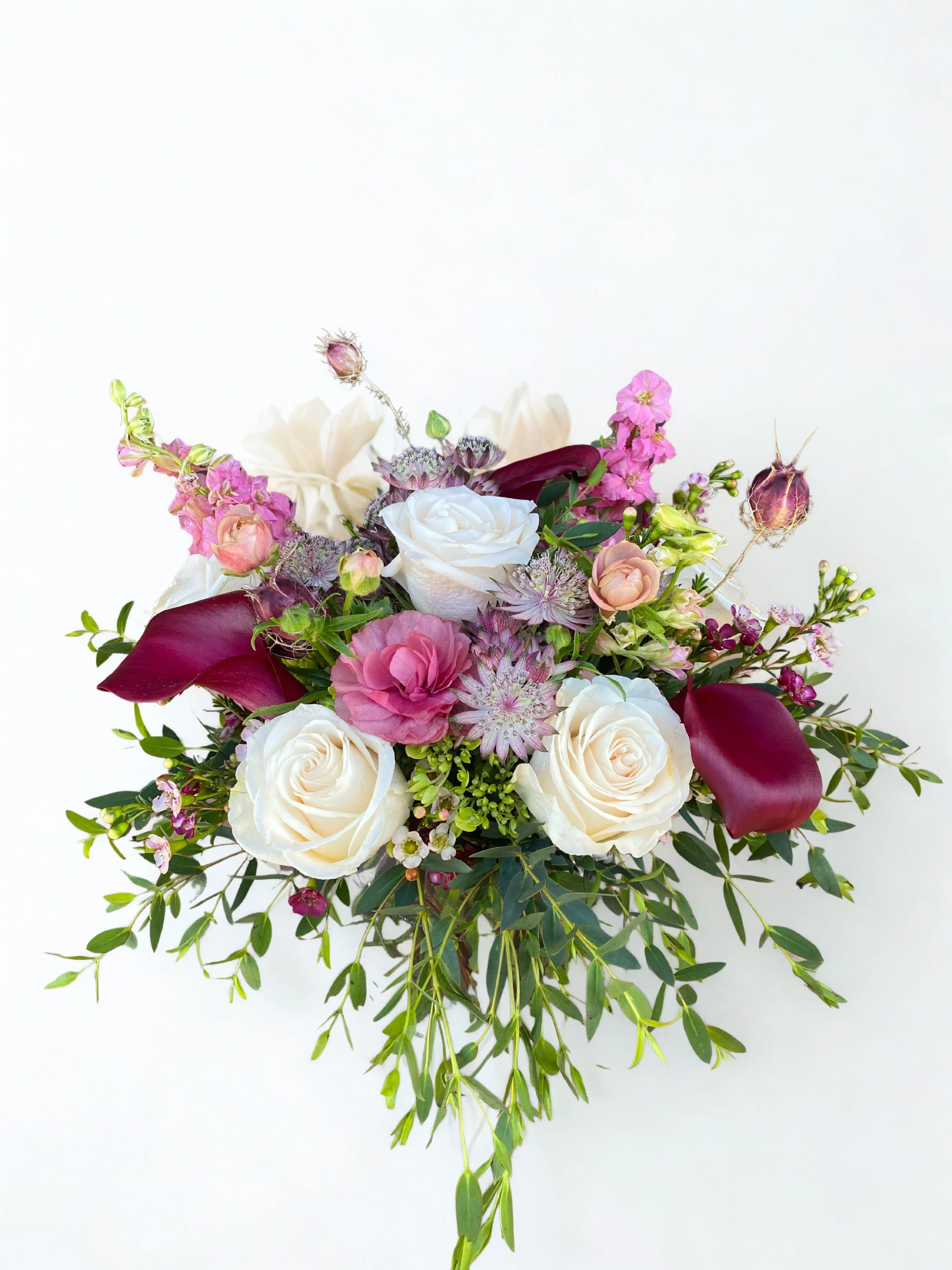 A bouquet of flowers including white roses, pink roses, pink and purple lilies, dark burgundy calla lilies, and various greenery against a white background.