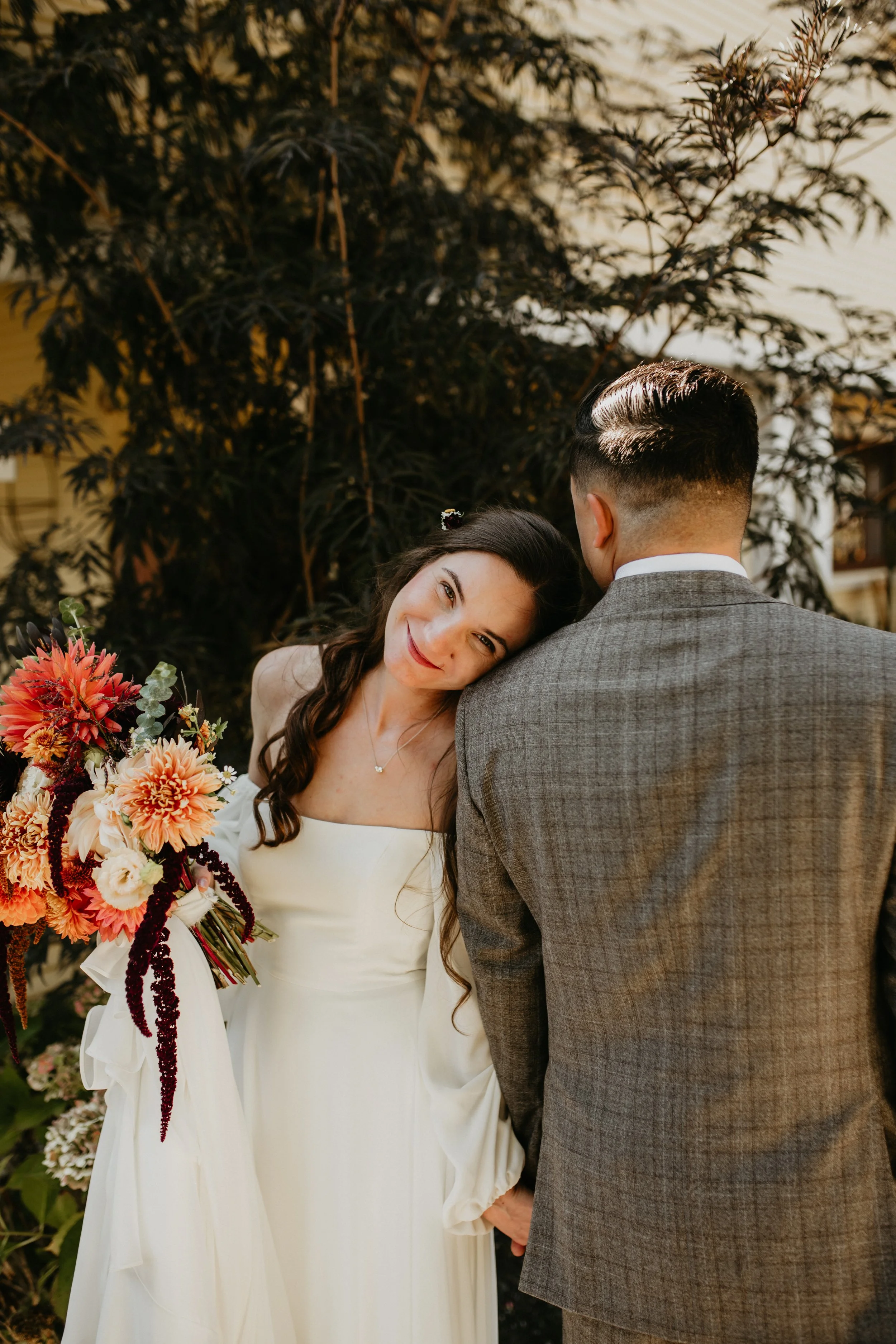 A bride with long dark hair and a white dress smiling and resting her head on her groom's shoulder during a wedding outdoors at sunset, holding a large bouquet of colorful flowers.