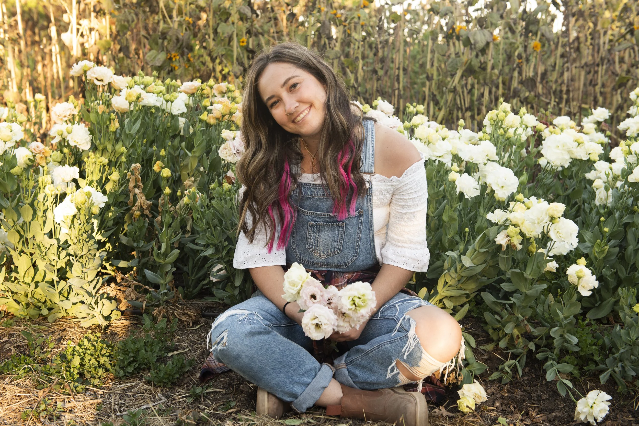 A young woman sitting on the ground in a flower garden, holding a bouquet of flowers, smiling, with pink highlights in her hair, wearing a white off-the-shoulder top, denim overalls, ripped jeans, and brown boots.