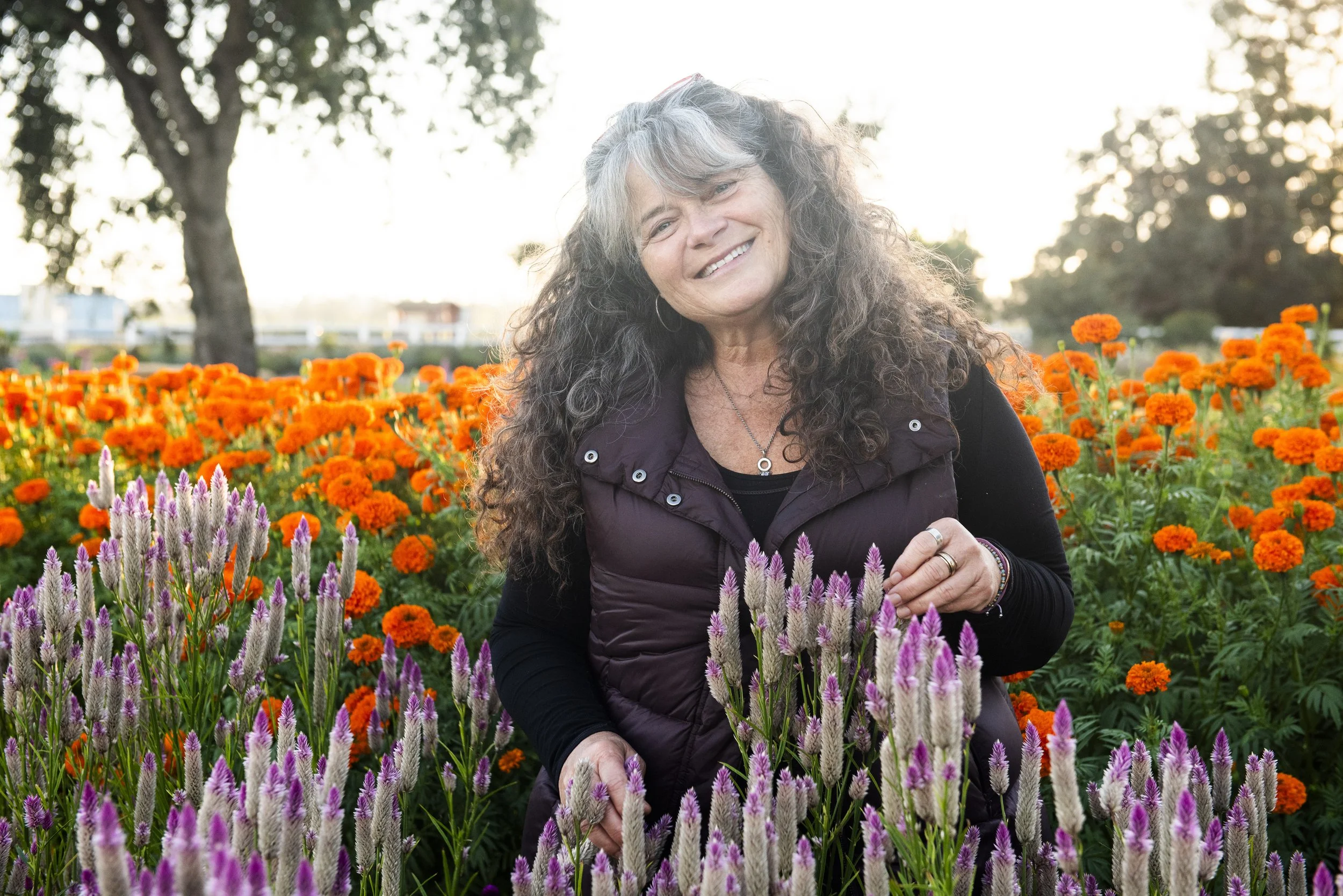 A smiling woman with long, curly gray hair stands among orange and pink flowers in a garden during sunset.
