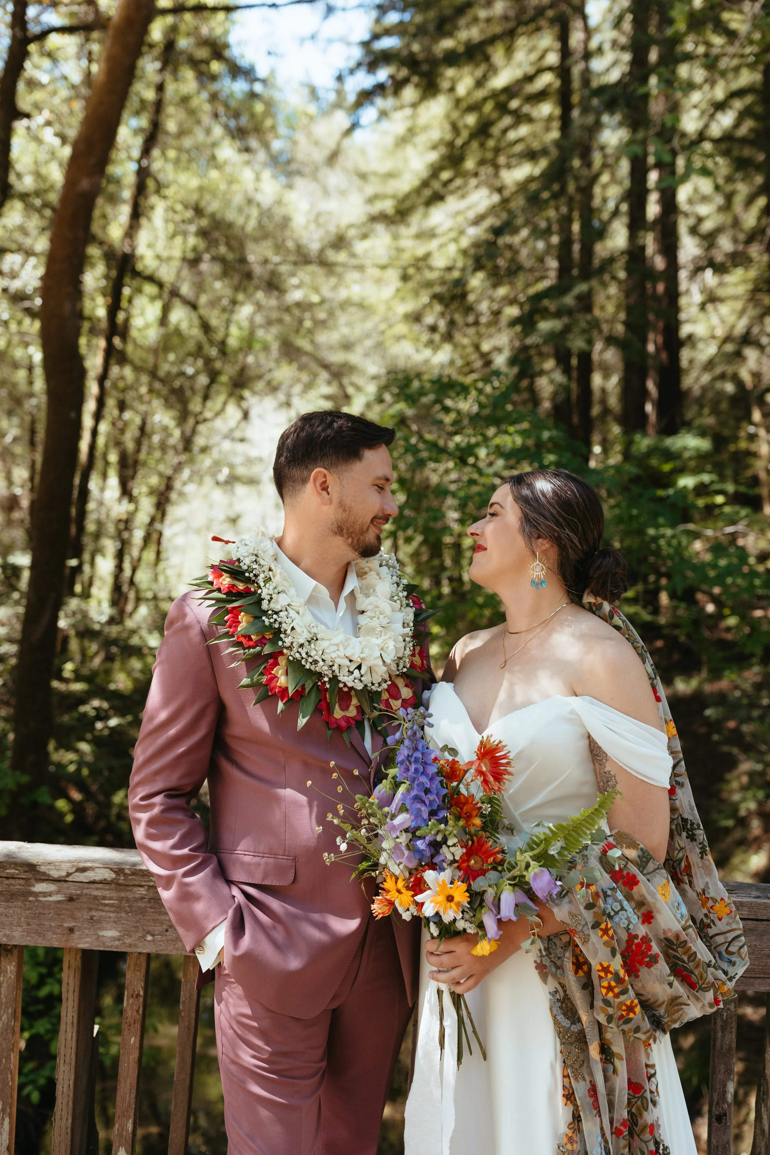 A bride and groom stand facing each other on a wooden bridge in a wooded area, smiling and holding a bouquet of flowers. The groom wears a mauve suit and a floral garland around his neck, while the bride wears a white off-shoulder dress and a patterned shawl.