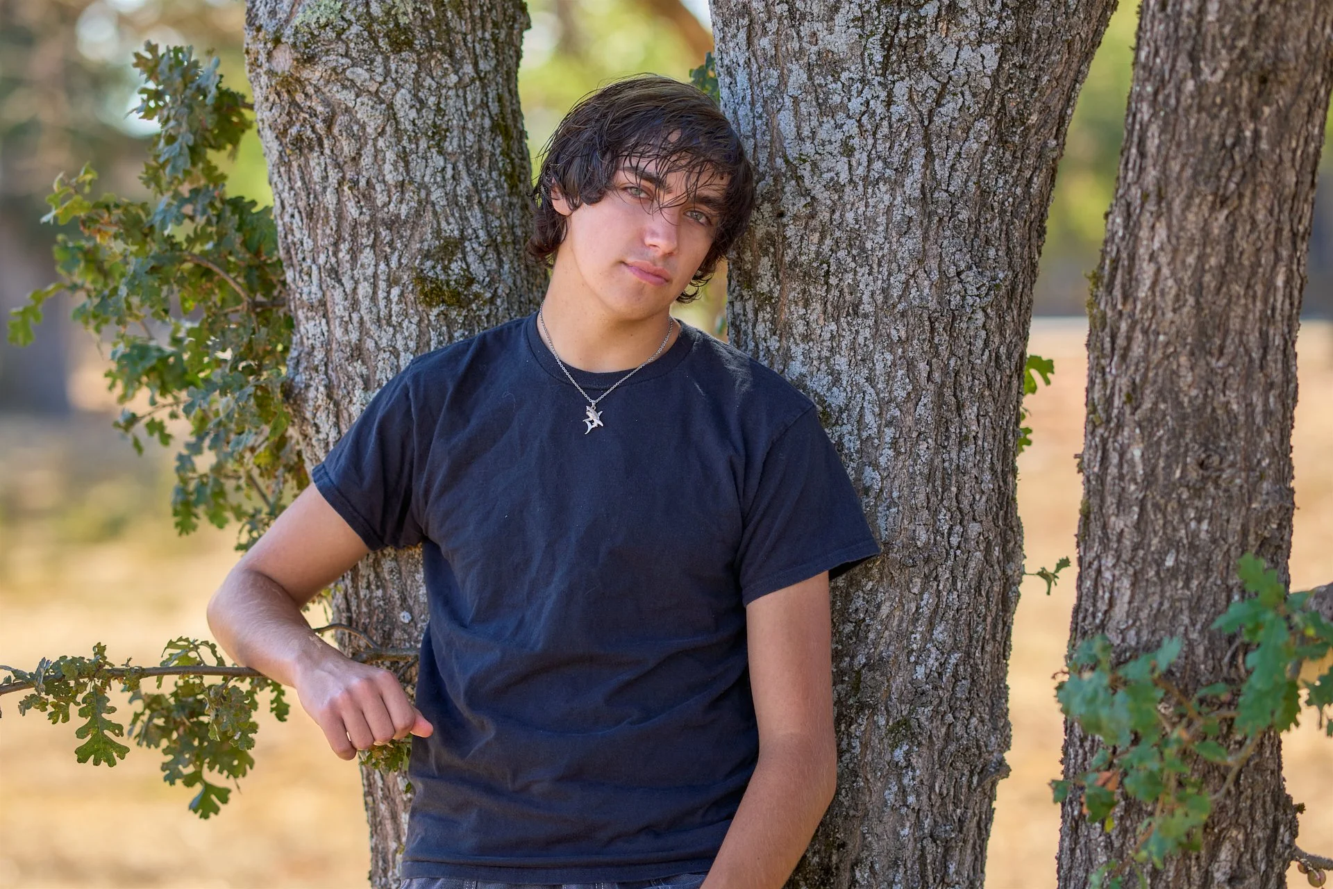 A young man with dark, wavy hair and light skin leaning against a tree outdoors, looking at the camera with a neutral expression. He is wearing a black T-shirt and a necklace with a star-shaped pendant.