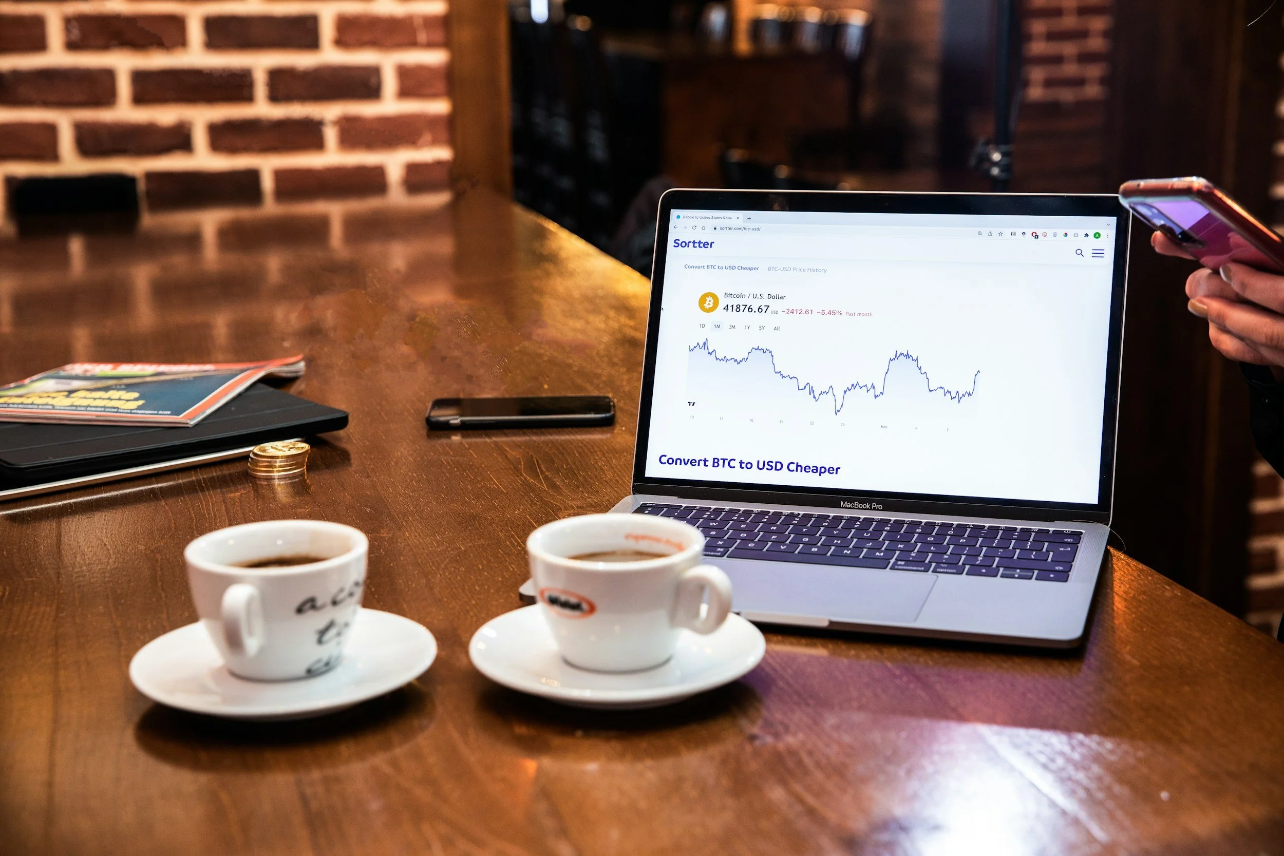 A laptop displaying a cryptocurrency chart on a wooden table with two cups of coffee, a smartphone, a stack of magazines, a pile of coins, and a closed notebook.