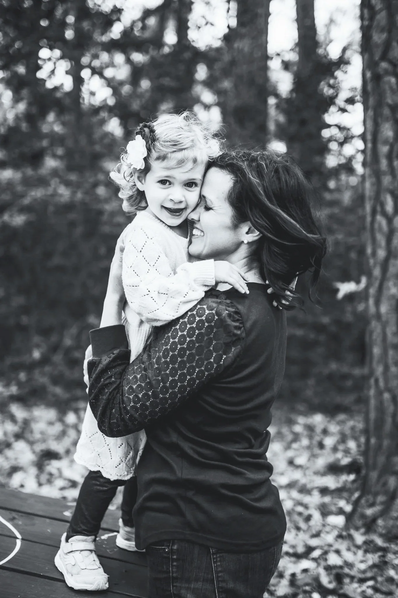 A woman and a young girl, possibly mother and daughter, sharing a hug outdoors in a wooded area. The girl is smiling and the woman is smiling with her eyes closed.