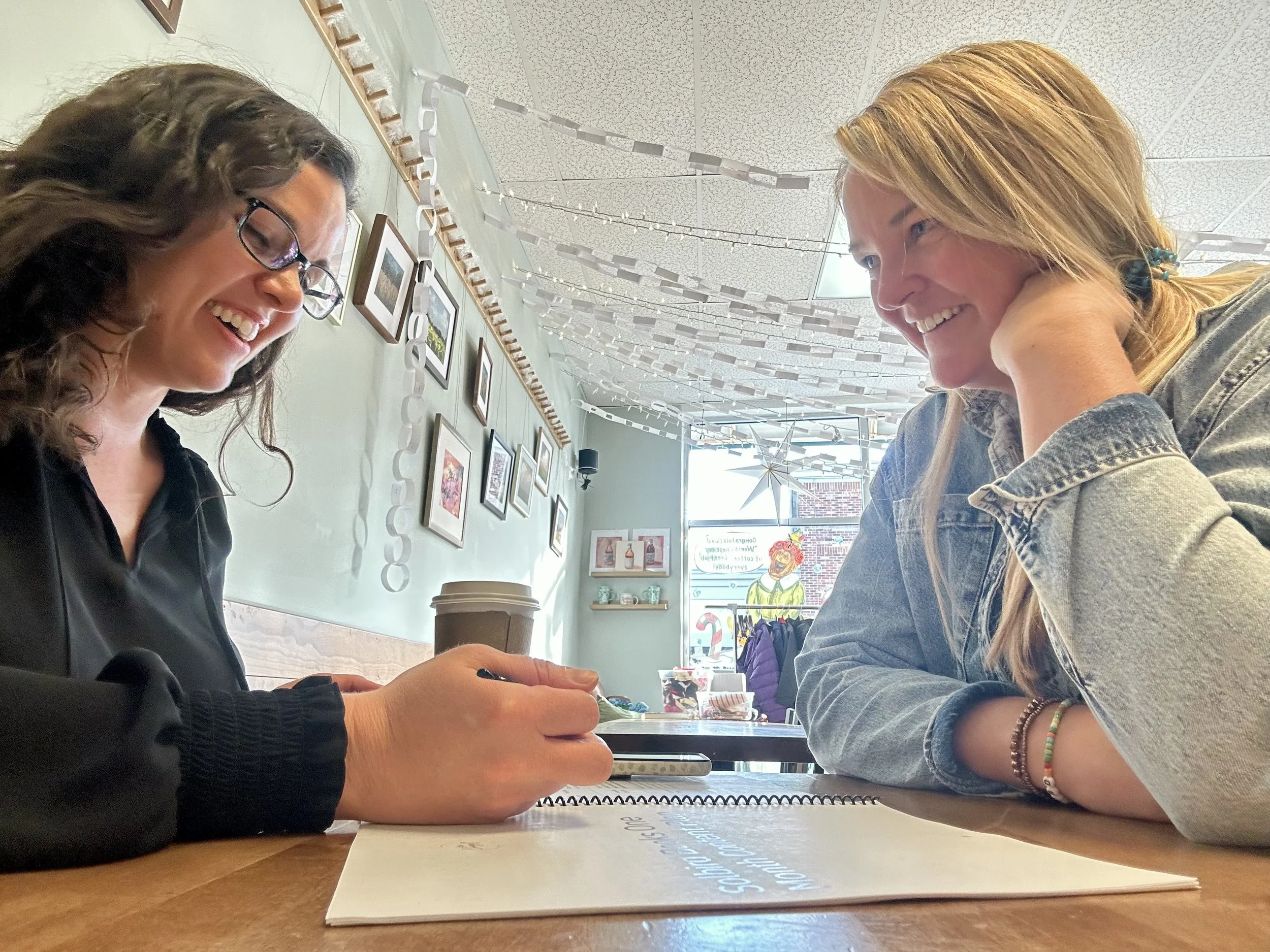 Two women sitting at a table, smiling and talking in a cafe decorated with pictures and hanging paper chains; one woman wears glasses and a black top, the other has blonde hair and a denim jacket.