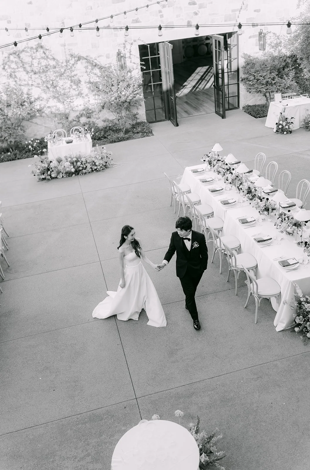 A bride and groom dancing at their wedding reception. The area is decorated with flowers, string lights, and a long table set for guests.