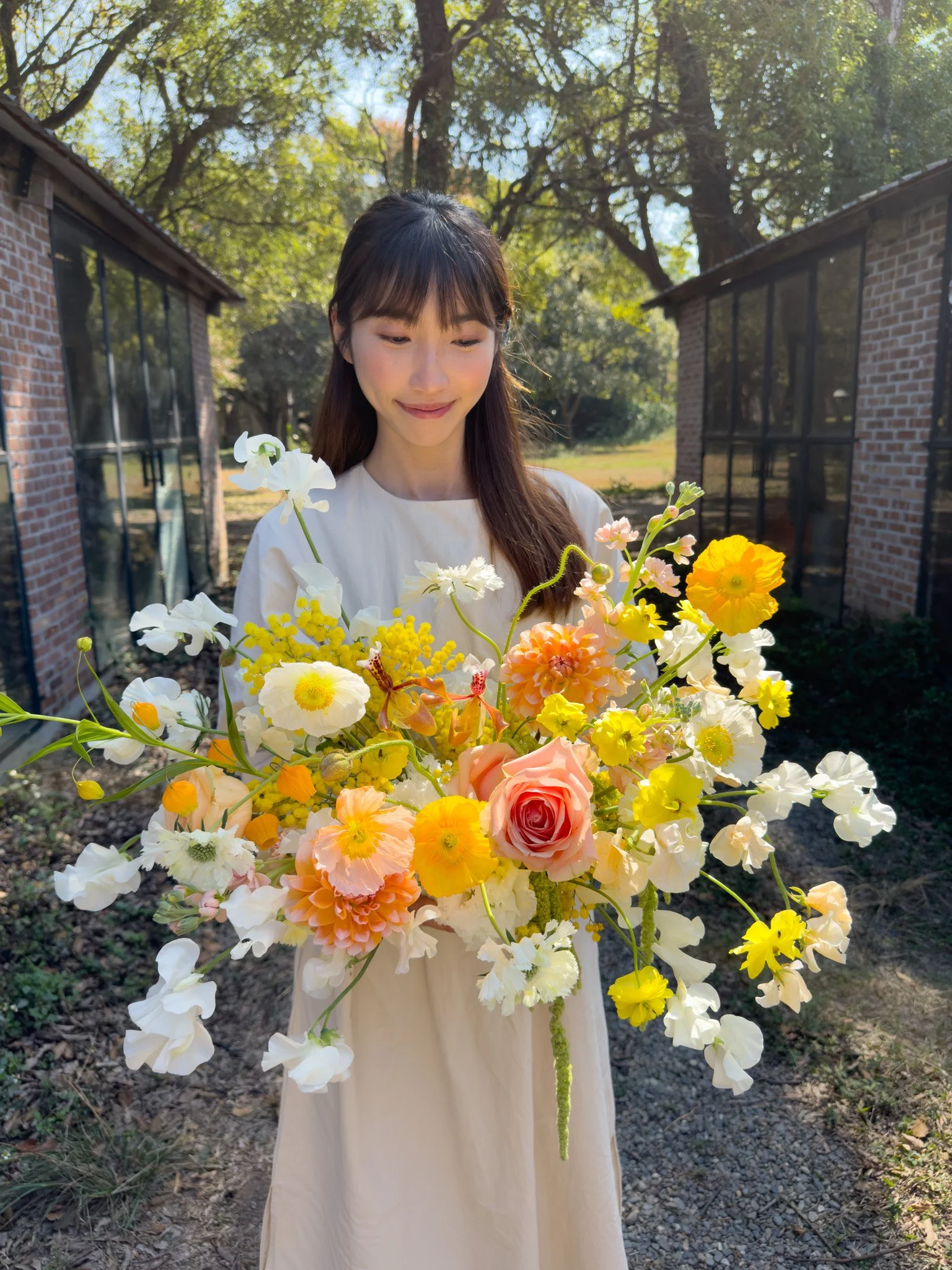 A young woman in a white dress holding a large colorful bouquet of flowers outdoors on a sunny day, with trees and brick buildings in the background.
