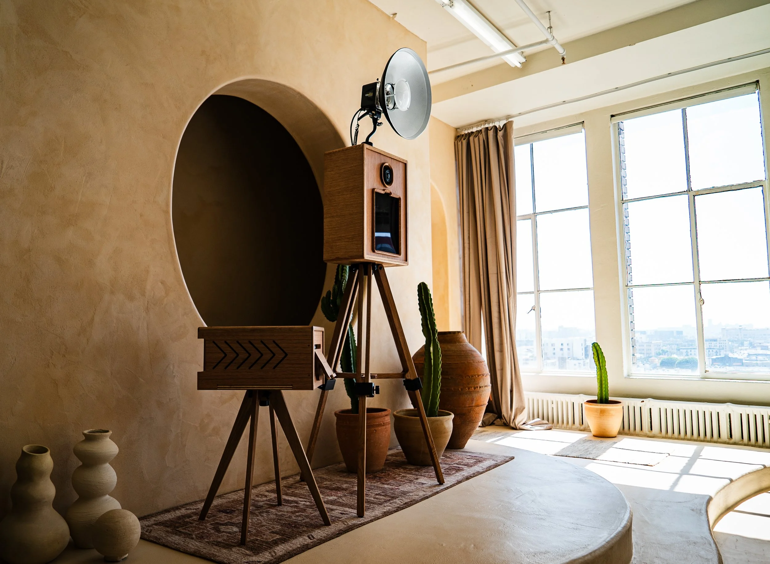 Interior of a room with large windows, curtains, and potted cacti, featuring a wooden tripod stand with a vintage camera, speaker, and a large light reflector.