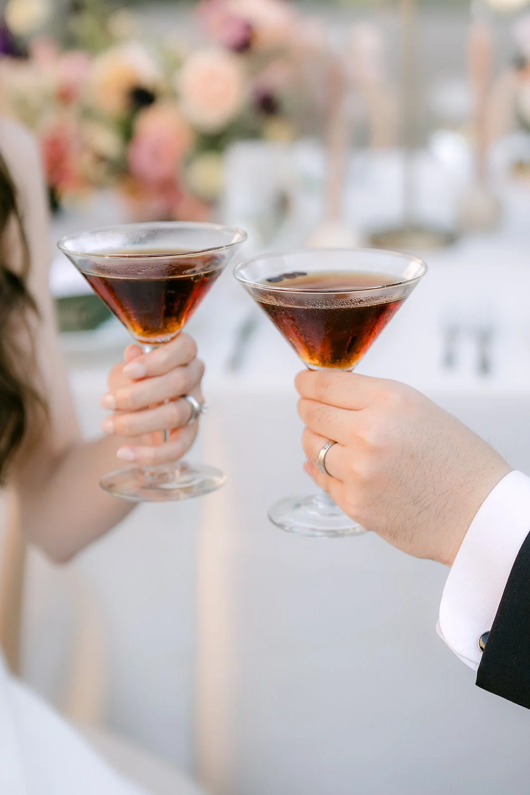Two people clink glasses filled with red drinks at a celebration or wedding reception, with floral decorations in the background.