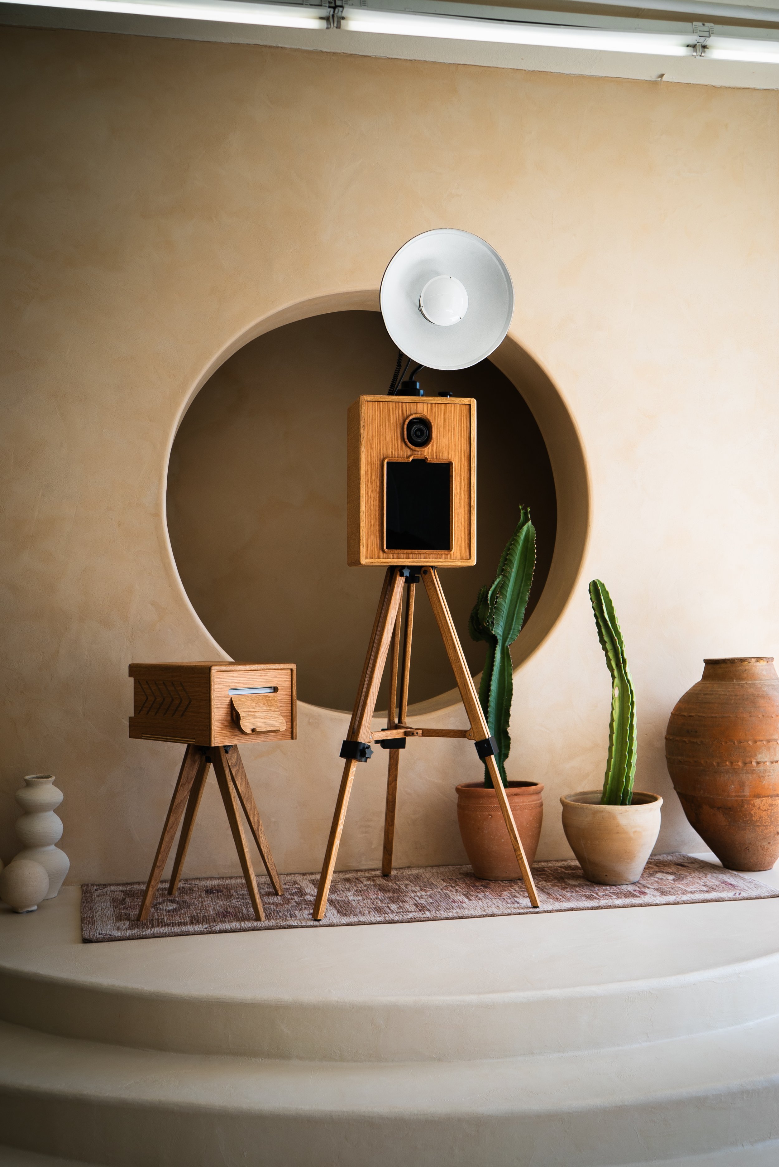 Indoor scene with a vintage wooden speaker on a tripod, a wooden side table, potted cacti, and decorative vessels against a beige wall with a circular recess.