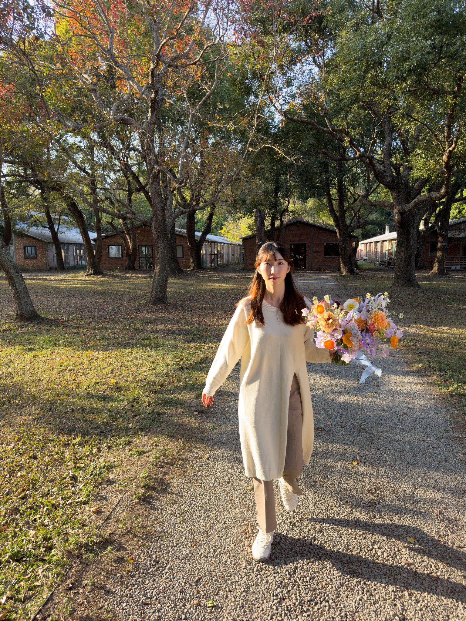 A young woman with long brown hair wearing a beige coat, beige pants, and white sneakers holding a large colorful bouquet of flowers, walking on a gravel path in a park with trees and small buildings in the background during late afternoon.