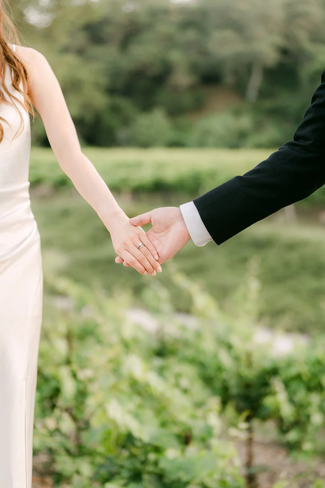 A woman in a white dress and a man in a black suit holding hands outdoors with greenery in the background.