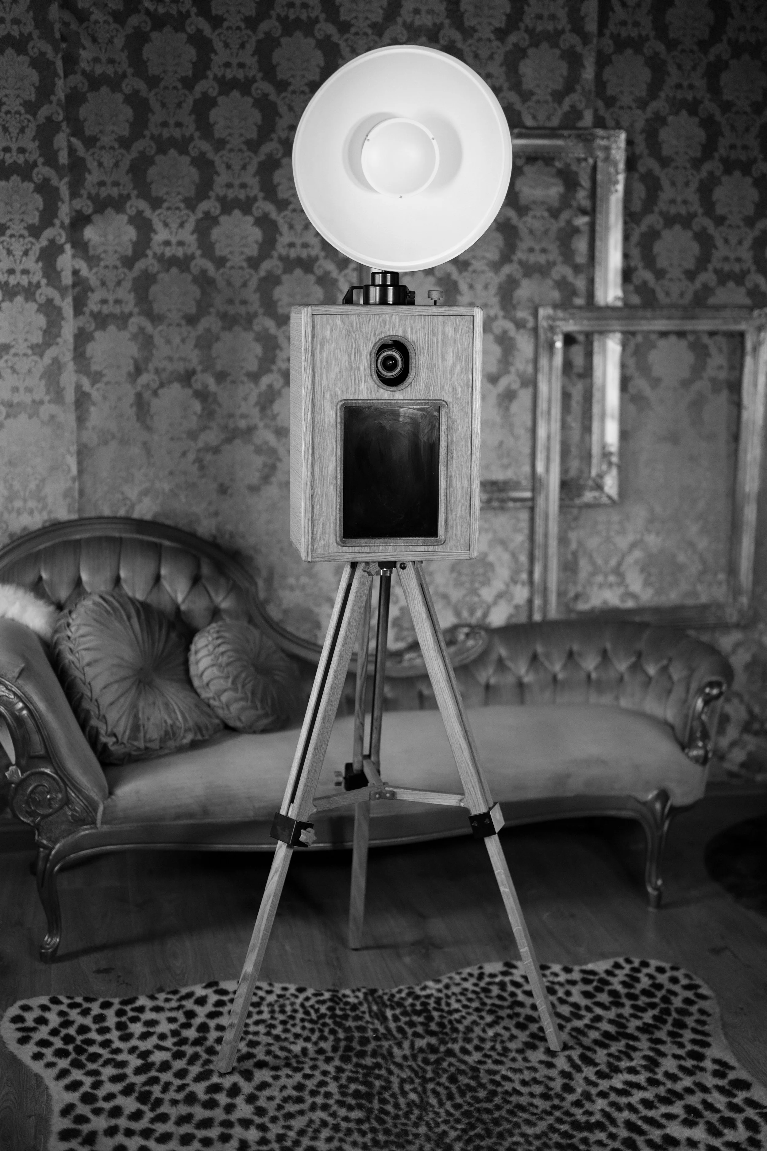 A vintage camera on a wooden tripod in a room with patterned wallpaper, velvet couches, ornate picture frames, and a leopard print rug.
