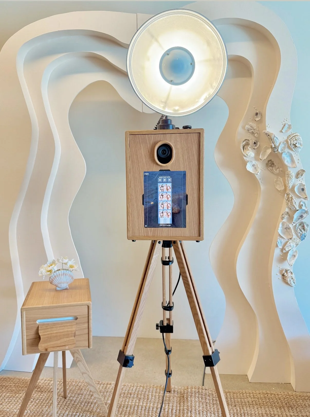 A photo booth with a large professional camera, a wooden box housing electronics, a studio light, a small wooden side table with a flower arrangement, set against an artistic wall with wave-like patterns and seashell decorations.