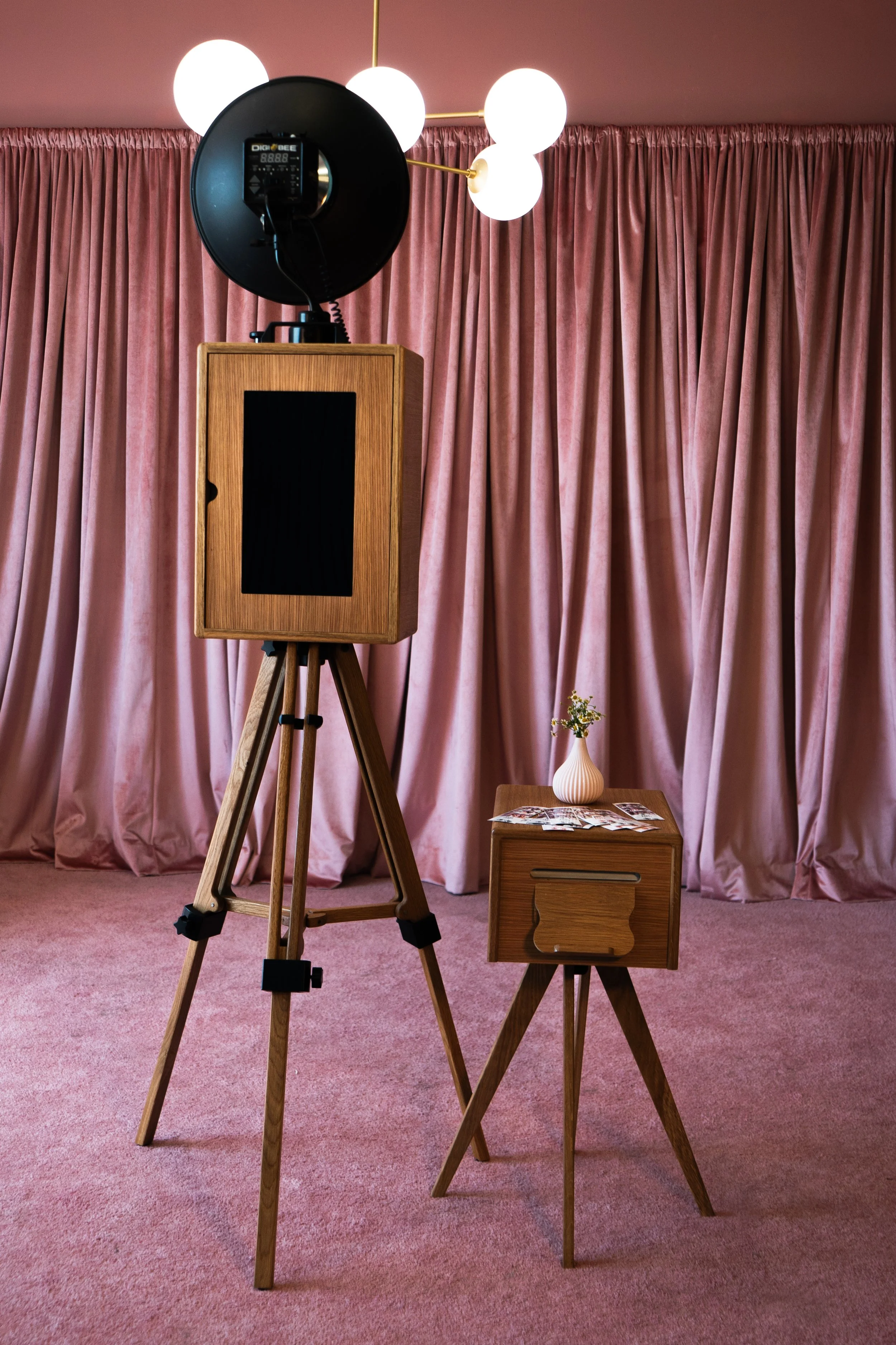Photography studio setup with a large vintage wooden camera on a tripod, a lamp with a black shade, pink curtains, a small wooden table with magazines, and a white vase with flowers.