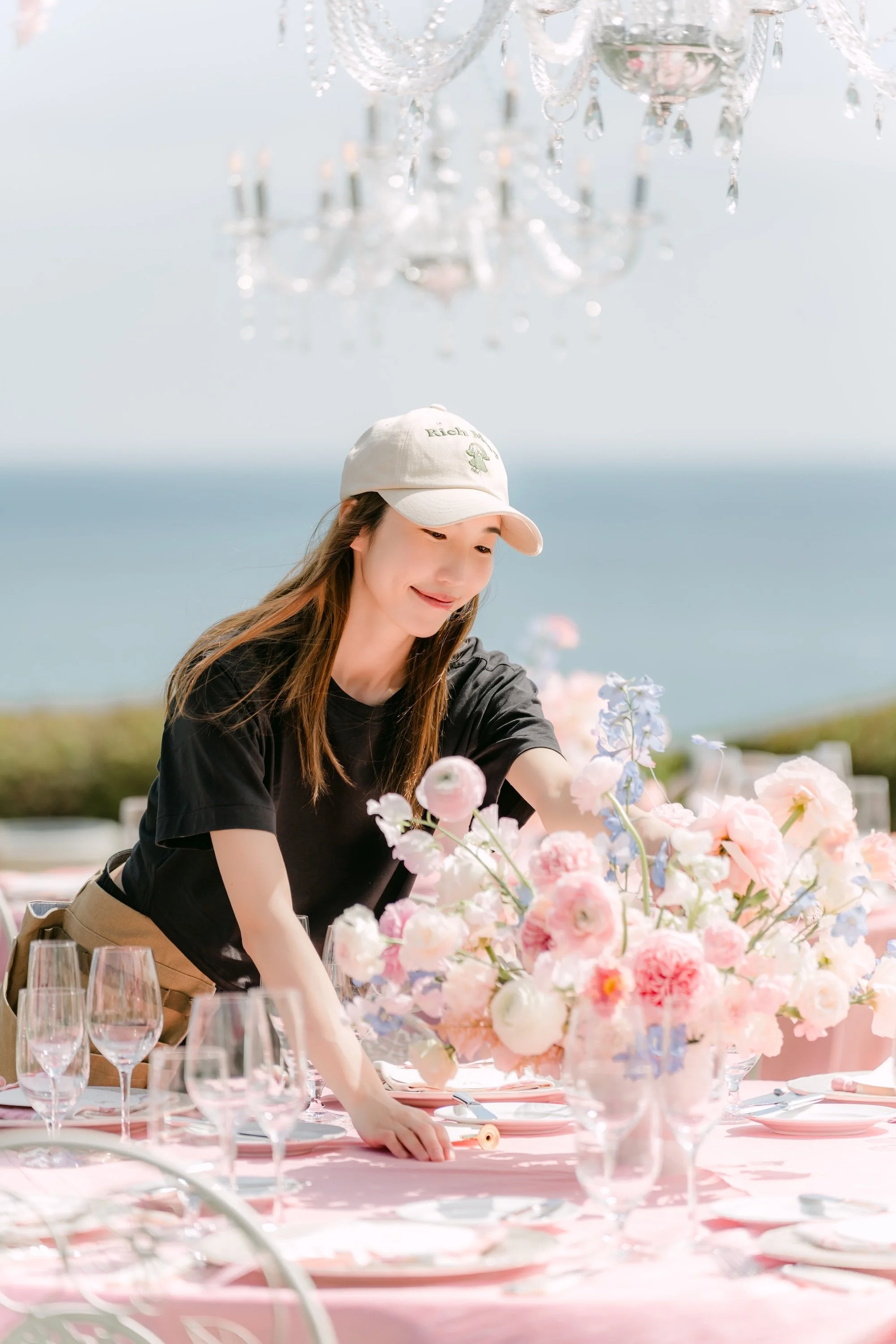 A woman decorating an outdoor table with pink flowers, with the ocean in the background.