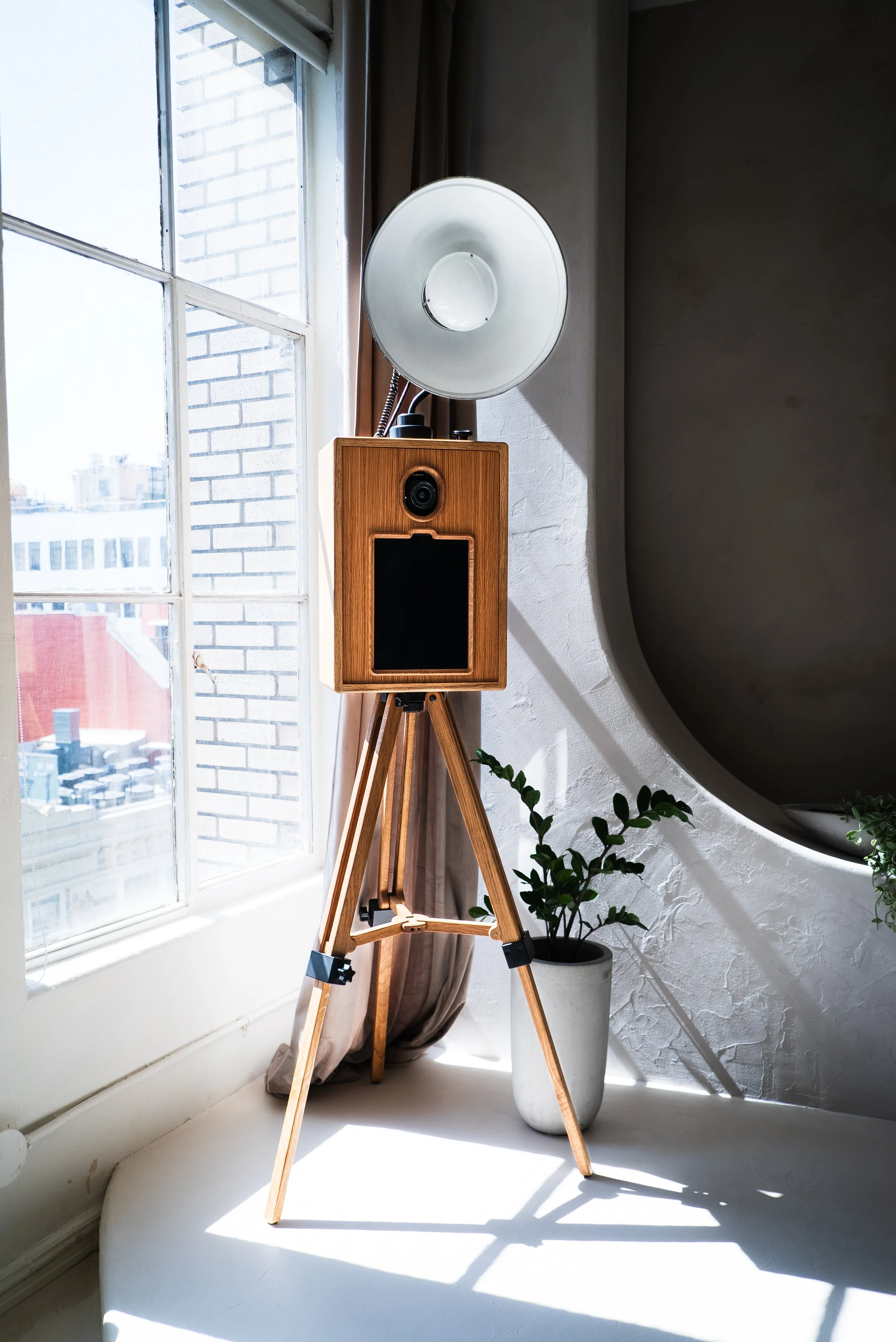 A wooden speaker with a black front panel, mounted on a tripod, positioned near a window with sunlight streaming in and a houseplant in a white pot beside it.