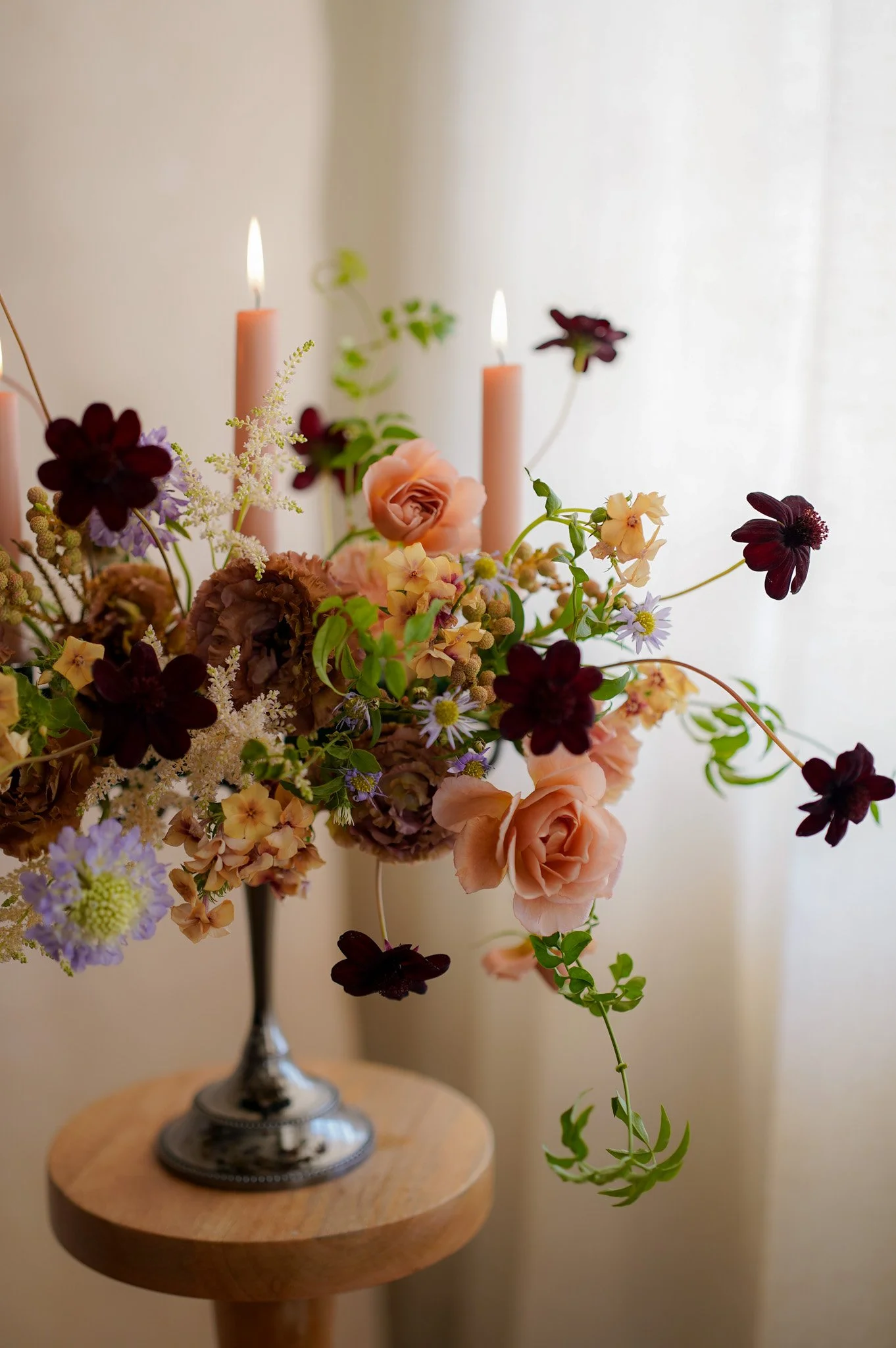 A floral arrangement with pink and dark purple flowers, set in a silver vase on a wooden table, with lit pink candles and a sheer curtain in the background.