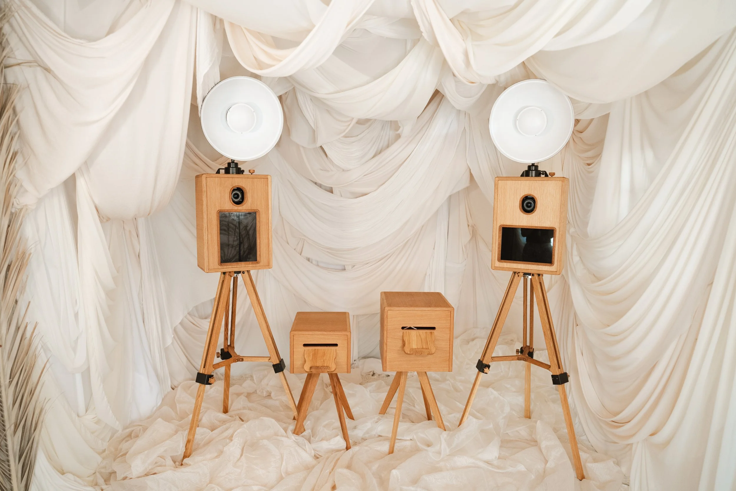 A photography setup with two large softbox lights mounted on wooden tripods, two wooden speaker boxes, and two wooden stools with pull-out drawers, all arranged in a white fabric-draped backdrop.